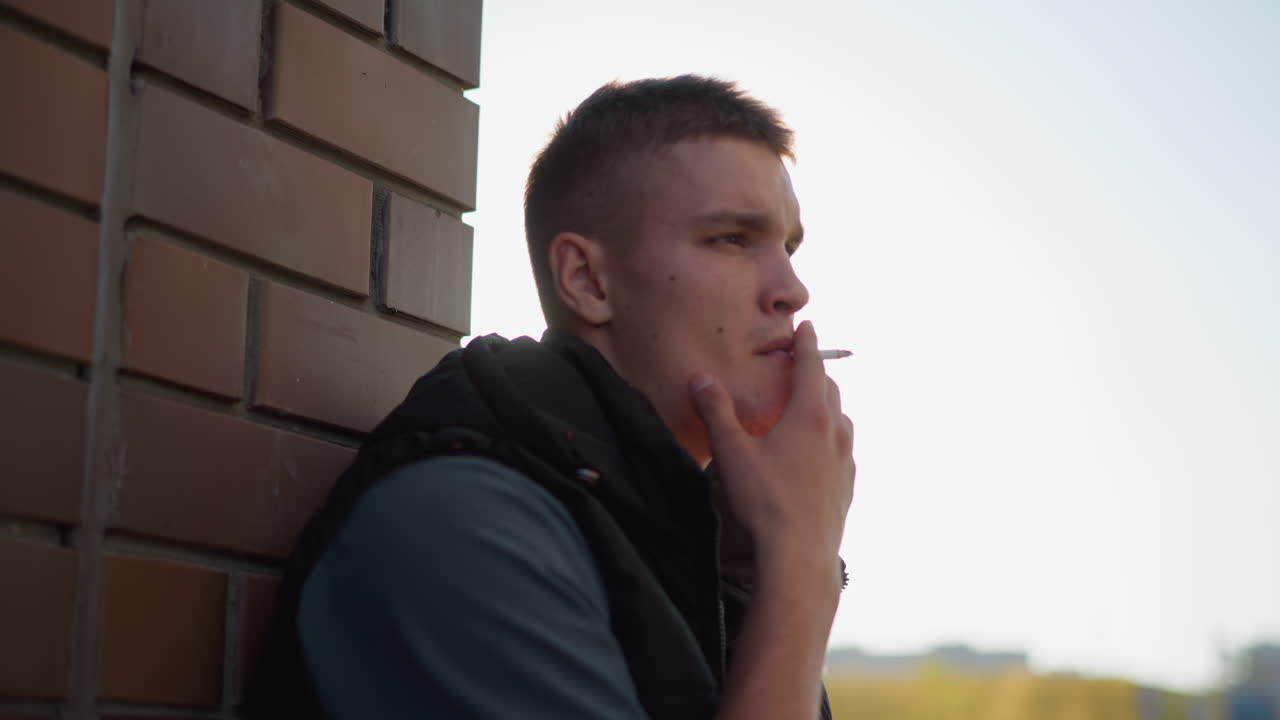 man leans back against brick wall holding cigarette close to lips while exhaling smoke, bathed in soft natural light with calm expression as he gazes into distance during daytime
