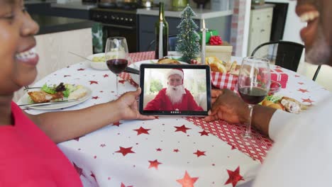 pareja afroamericana con vino usando tableta para la videollamada de navidad con feliz santa en la pantalla