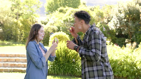 Feliz-Pareja-Diversa-Soplando-Burbujas-En-Un-Jardín-Soleado,-En-Cámara-Lenta