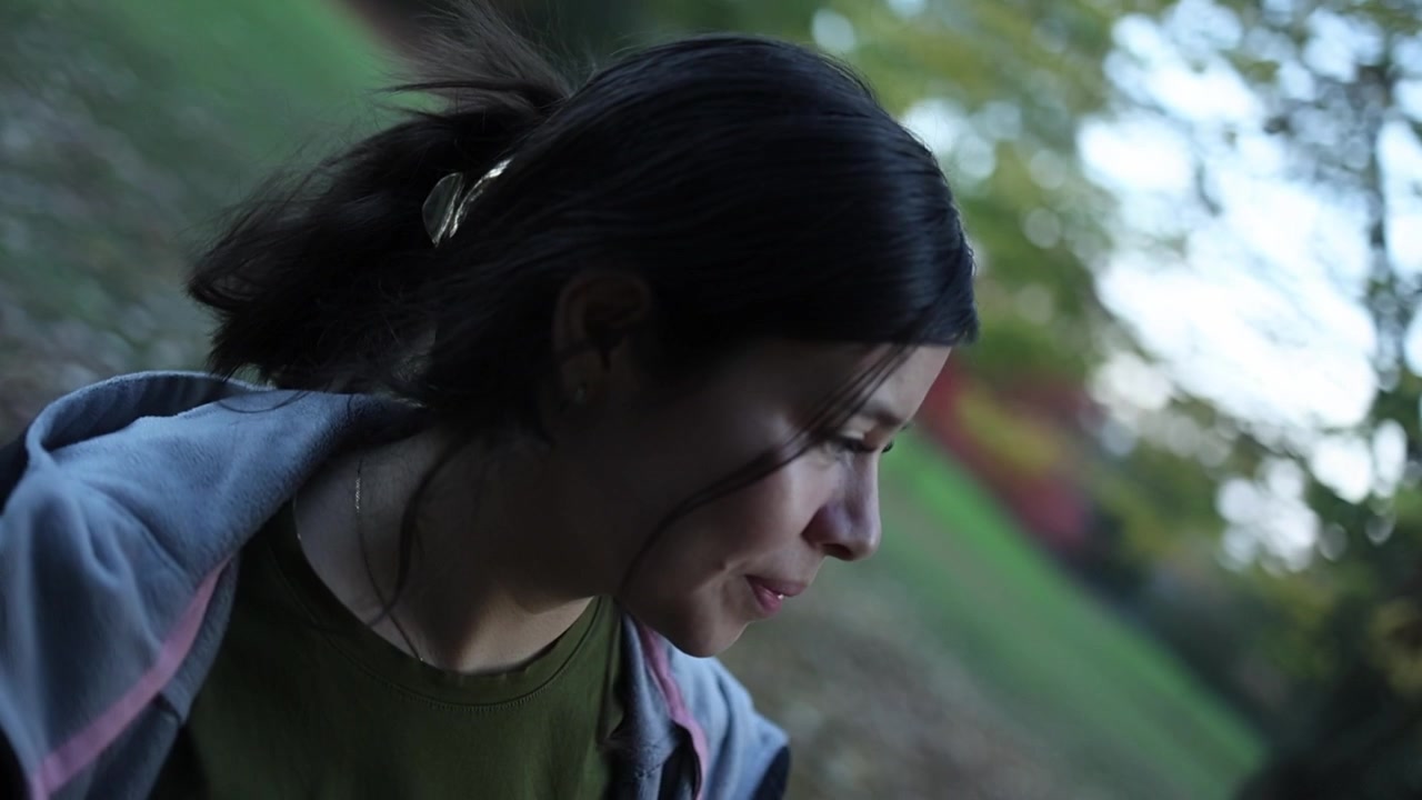 一位留著深色頭髮、戴著花形髮夾的年輕女子，若有所思地走過綠葉公園，周圍環繞著柔和的綠植和黃昏的光線，以電影感、親密的特寫鏡頭呈現。