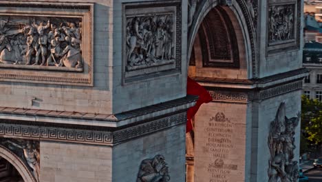 aerial drone close up footage flying around city gates of paris that has a large french flag waving in the middle of it in slow motion during sunset or sunrise. the stone gates have sculptures on it.