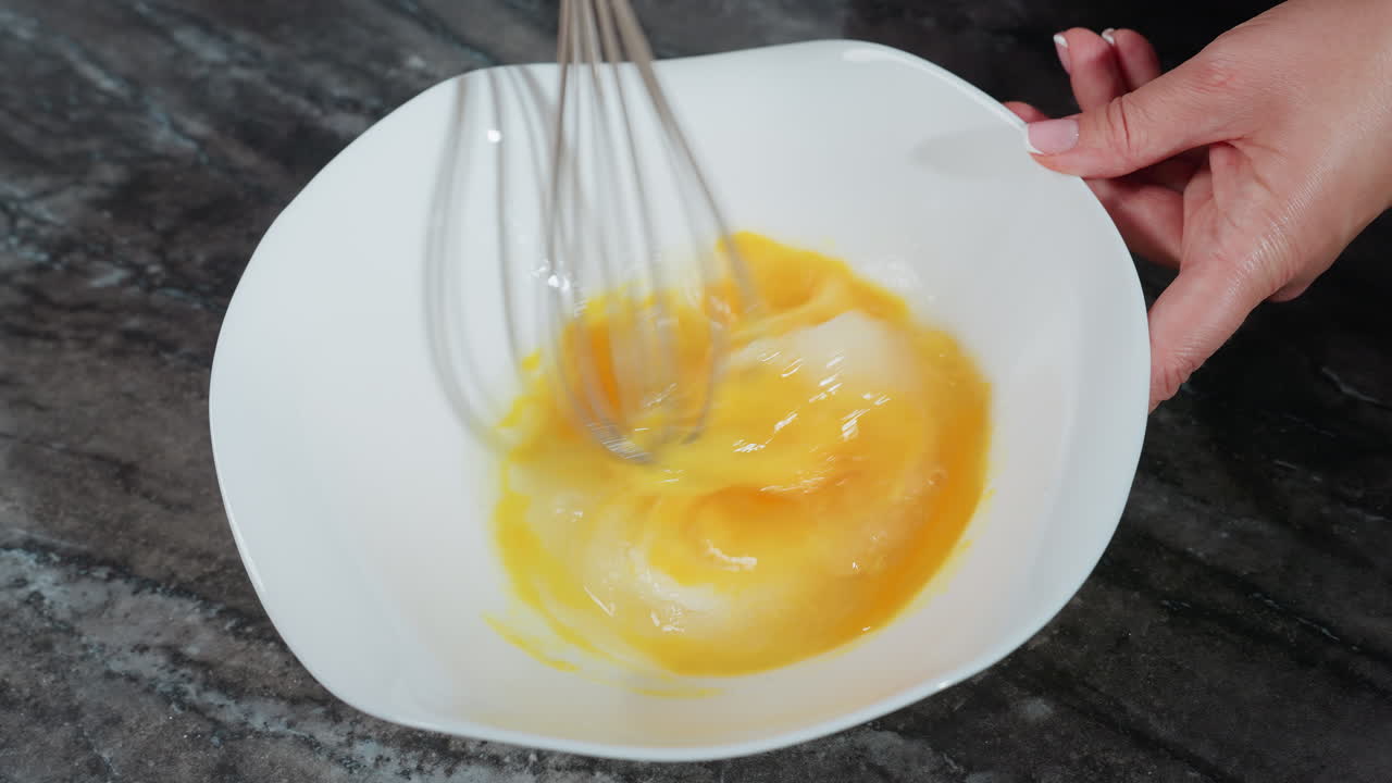 primo piano dello chef che sbatte vigorosamente le uova in una ciotola bianca, mescolando tuorli e zucchero per creare una consistenza liscia, tenendo la ciotola ferma su un bancone scuro
