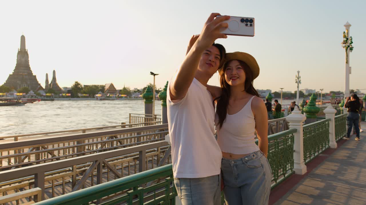 Coppia sorridente posa per un selfie vicino a un tempio sul fiume, illuminazione dell'ora d'oro, atmosfera rilassata da vacanza