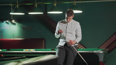 young player in white shirt and grey trousers sits on green pool table, holding cue stick with a focused expression. another pool table is visible in the background under dim lighting