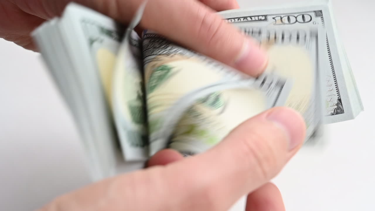 A man's hands quickly count a stack of one hundred dollar bills against a light background