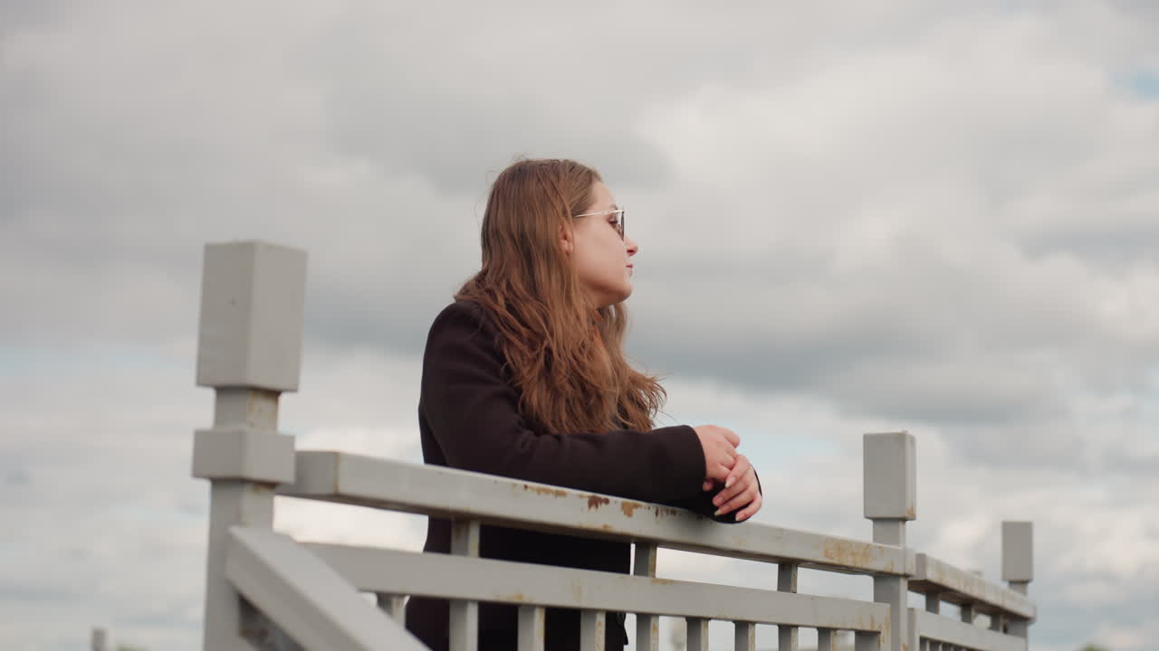 White American lady with long brown hair and sunglasses walks towards guard rail under grey sky, showing calmness and thoughtful mood while gentle daylight highlights her natural expression and style