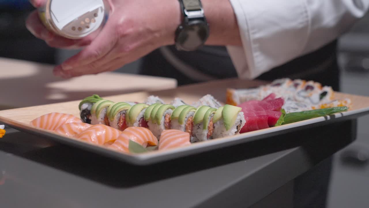 A sliding shot captures a sushi chef sprinkling pepper flakes onto an avocado sushi roll, enhancing flavor and presentation. Perfect for culinary, sushi preparation, and professional kitchen content.