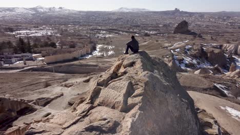 cappadocia adventure: solo tourist hiker sits atop mountain cave dwelling enjoying view towards göreme and uchisar castle on horizon