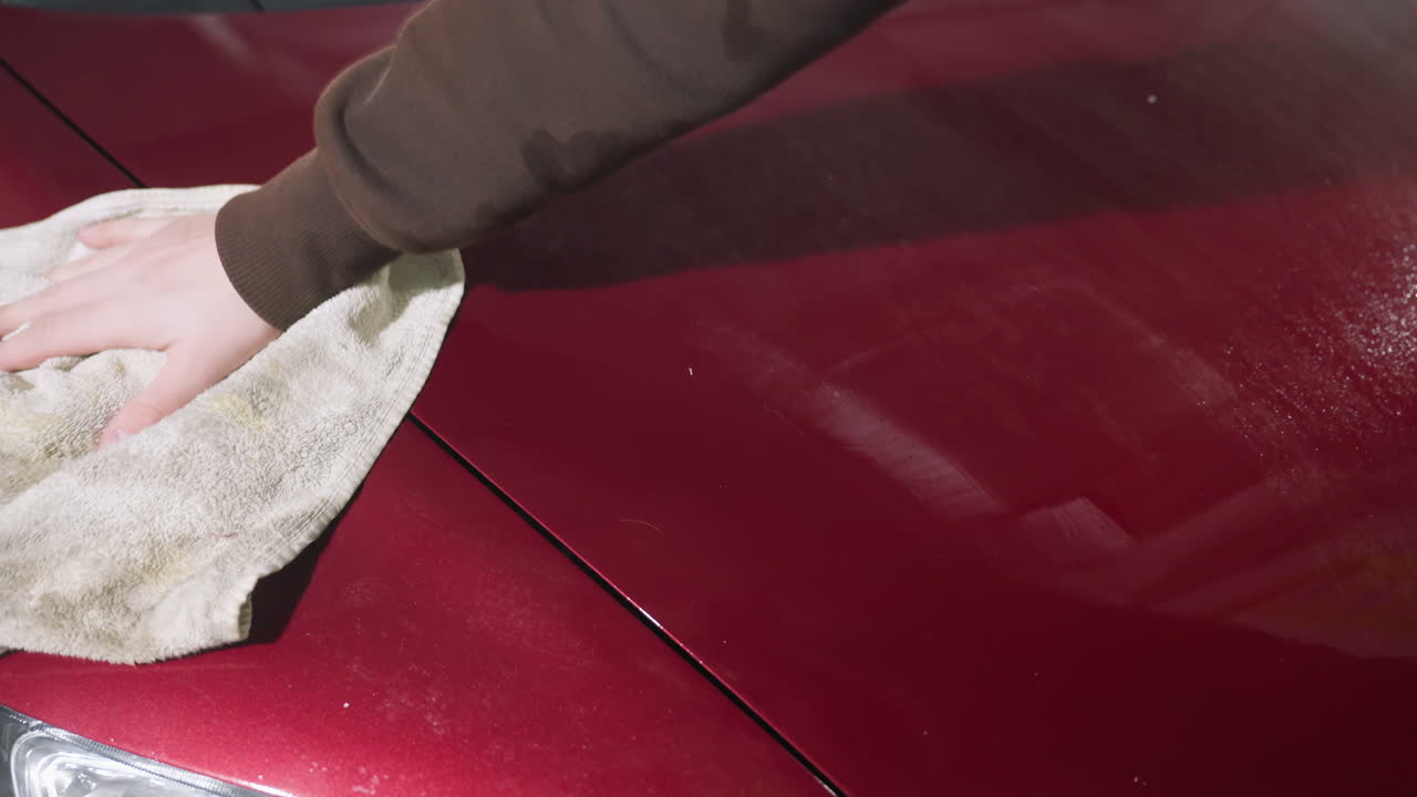 close up of person wearing brown hoodie wiping water off shiny red car bonnet and headlamp with soft cloth during car detailing session under bright indoor lighting reflecting on surface