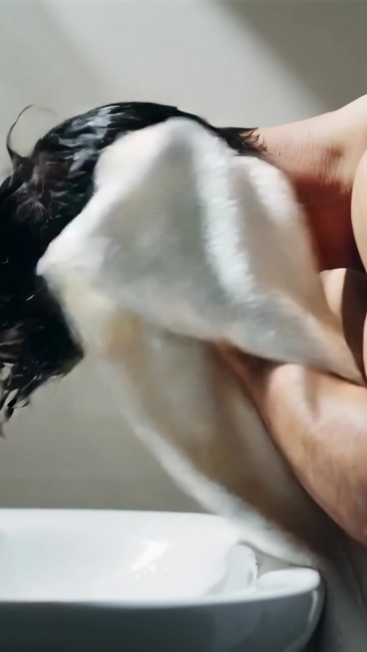 Video vertical: Hombre sin camiseta secándose el pelo mojado con una toalla blanca en el lavabo, con el grifo visible.