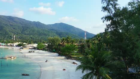 Beautiful-leaves-of-palm-trees-hanging-over-white-sandy-beach-washed-by-calm-turquoise-lagoon-at-sunrise-with-bright-sky-in-Thailand