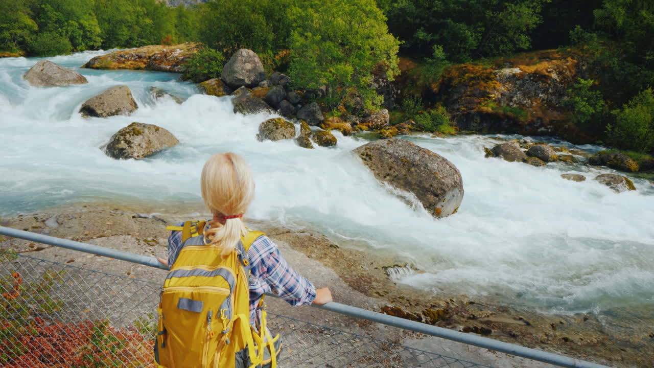 una mujer joven parada junto a la cerca mira el agua de deshielo del río de montaña escarpada que corre desde el briksda