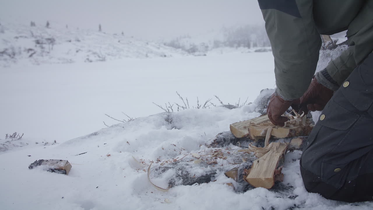 Hands creating wood shavings from logs on snow-covered ground in a foggy, frozen wilderness setting.