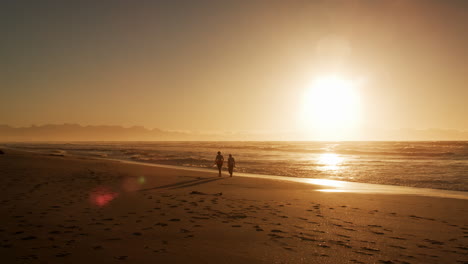 two silhouetted figures walk along beach by waves at sunset