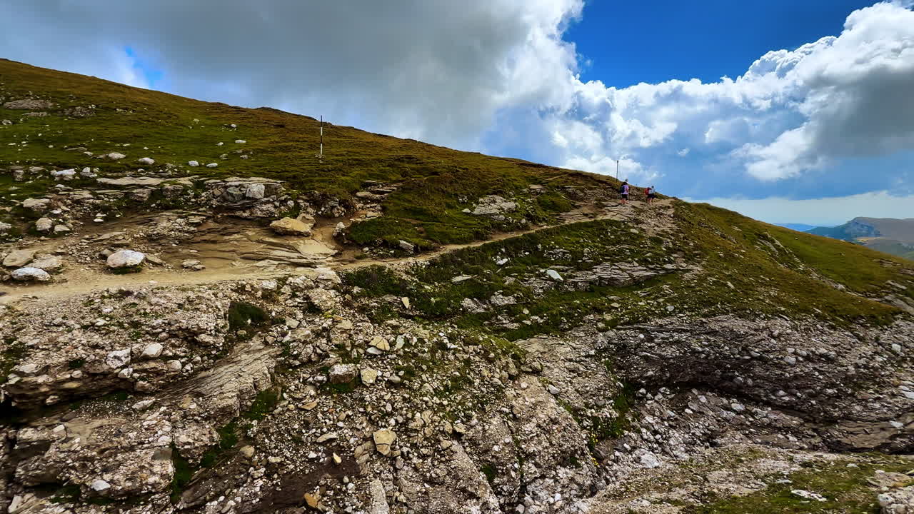 Escursionisti camminano lungo un sentiero roccioso in un paesaggio montano in una giornata di sole. Due persone percorrono un sentiero roccioso su una collina verde sotto un cielo blu con nuvole