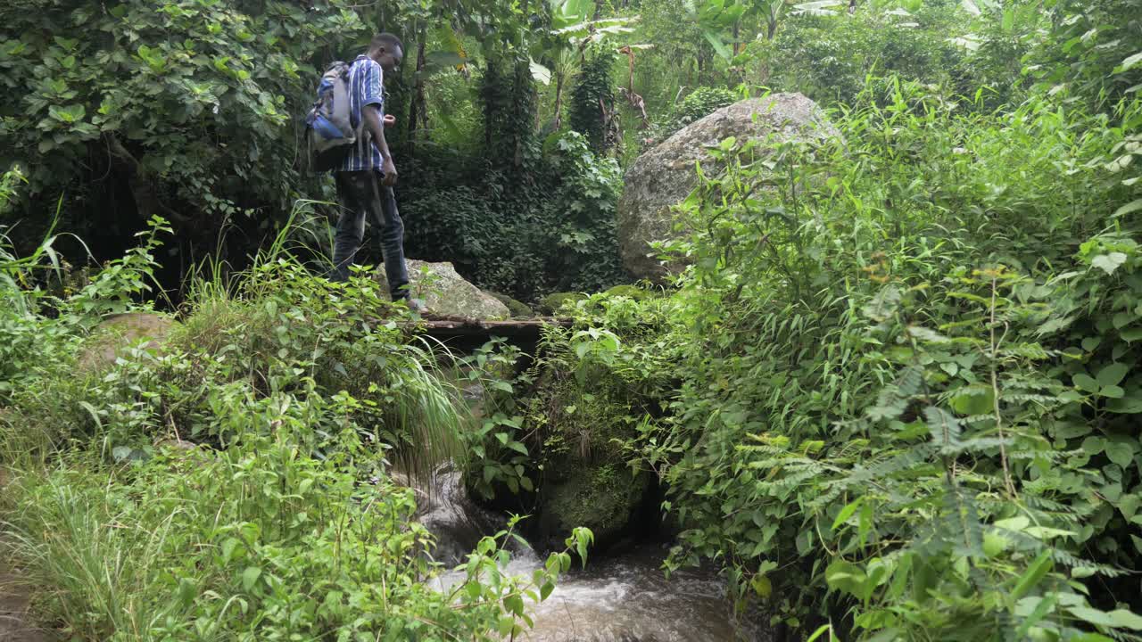 열대 동부 아프리카 정글의 개울을 가로지르는 작은 나무 다리를 건너는 아프리카 남자의 슬로우 모션 샷