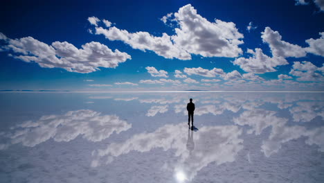man standing on a reflective salt flat under a vast blue sky with clouds