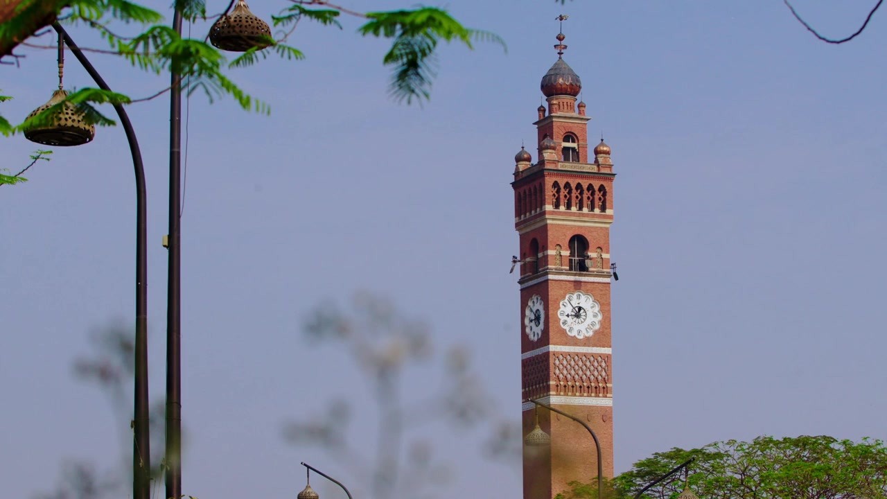 Ampia veduta panoramica dell'iconica Torre dell'Orologio di Husainabad a Lucknow, illuminata contro il cielo notturno. L'inquadratura cattura l'intera altezza della torre con i suoi dettagli ornati in stile gotico, che brillano