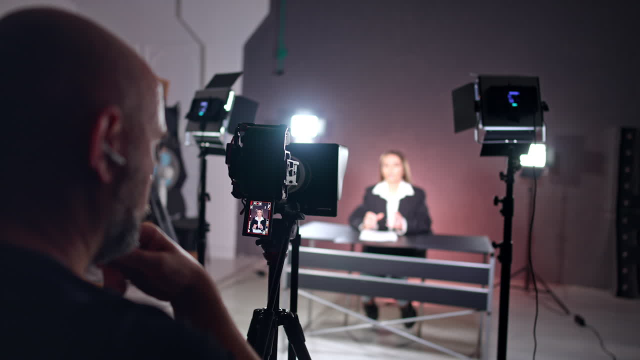 Backstage of the photo studio work. Male cameraman sits at camera recoding video of a female reporter.