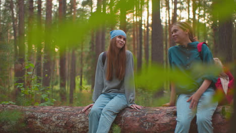 two friends on a fallen tree in a peaceful forest, the one wearing a blue bandana smiles joyfully, while her friend in green rests her hand on the tree, both looking at each other with a smile