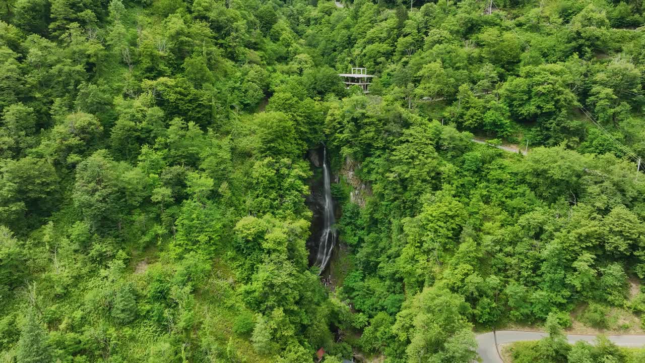 A tall and slender waterfall flows through the dense green mountains, surrounded by thick trees and hidden valleys