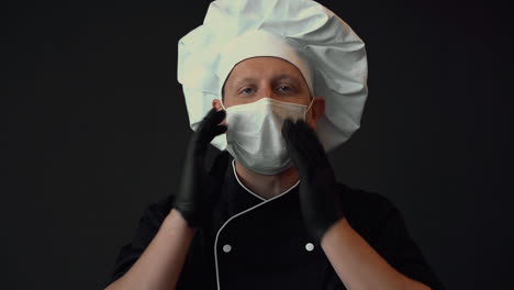 Chef-Masculino-Listo-Para-Cocinar,-Se-Pone-Mascarilla-Médica-Y-Hace-El-Gesto-De-Aprobación.-Retrato-Sobre-Fondo-Negro.