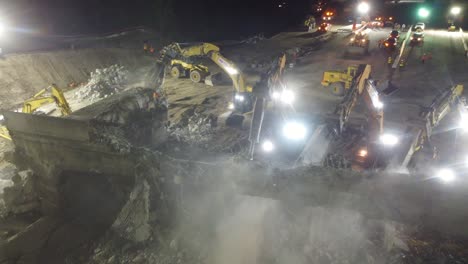 large group of heavy equipment demolishes a heavily reinforced concrete overpass in the night, overhead drone showcases dusty night environment