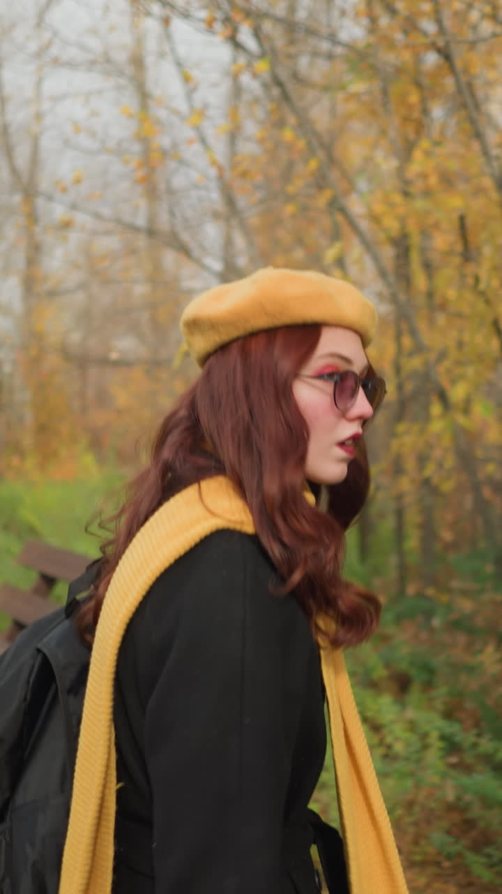 Seitenansicht einer Touristin in schwarzem Mantel, gelbem Schal und Baskenmütze, die einen hölzernen Pavillon in einem ruhigen Herbstpark betritt und ihr Haar richtet, während sie den Taschenriemen hält.