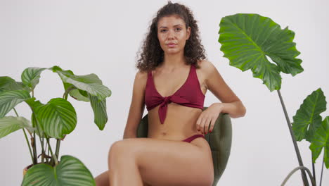 modelo bronceada y de cabello rizado oscuro posando en una silla con un traje de bikini rojo, rodeada de plantas, filmada en un estudio fotográfico