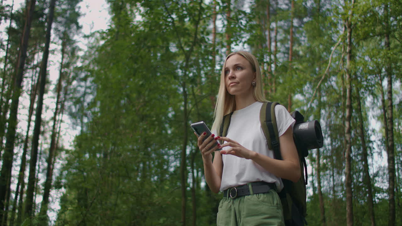 A young woman with a mobile phone walks through the forest traveling with a backpack in slow motion. Traveler in shorts in the woods looking for gps satellites