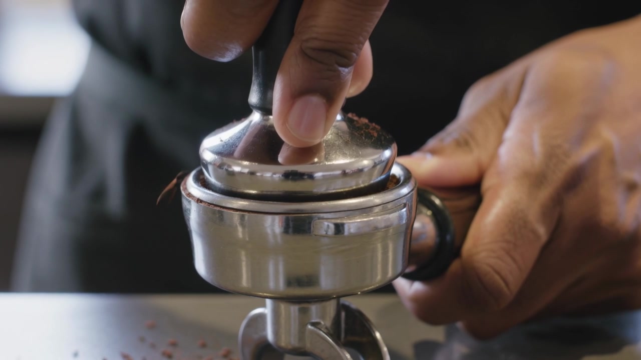 Barista de avental abaixando o compactador, estabilizando o porta-filtro e pressionando o pó para espresso em um café.