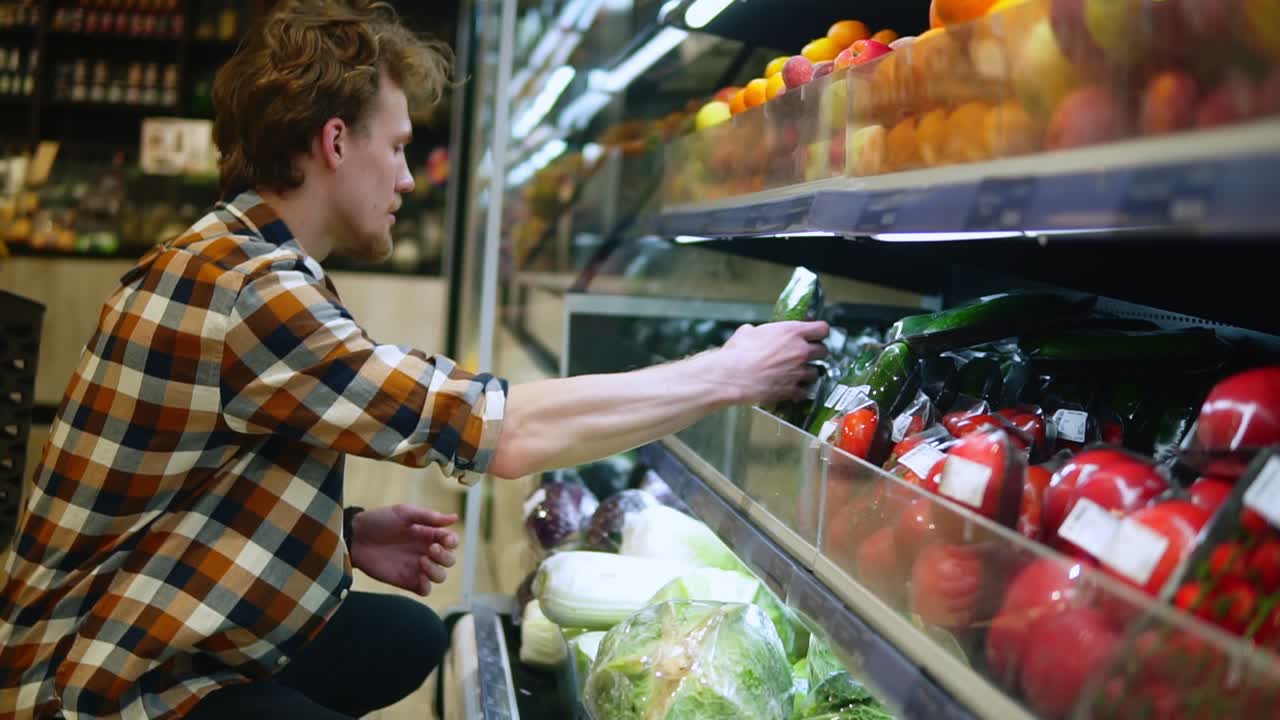 un hombre caucásico en camisa a cuadros comprando frutas y verduras en el departamento de productos de un supermercado de una tienda de comestibles. eligiendo pepinos frescos perfectos del estante inferior