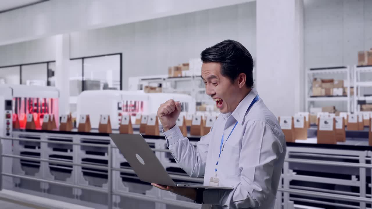 Side View Of An Asian Male Professional Worker Standing With His Laptop With Conveyor for Packaging Coffee Beans in Bags at Coffee Factory, He Raises His Fist Up With Screaming Goal