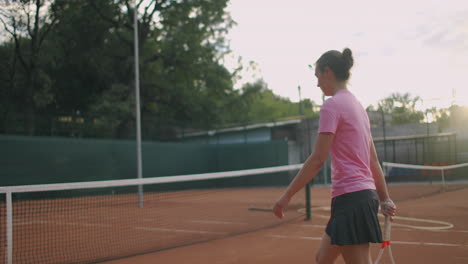 A-tired-brunette-female-tennis-player-walks-along-the-tennis-court-recuperating-and-concentrating.-Break-in-a-tennis-match.-Tennis-player-after-the-match-on-the-map-at-sunset-in-slow-motion.