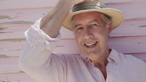 Portrait-of-happy-senior-caucasian-man-putting-on-hat-by-weathered-pink-wooden-wall,-in-slow-motion