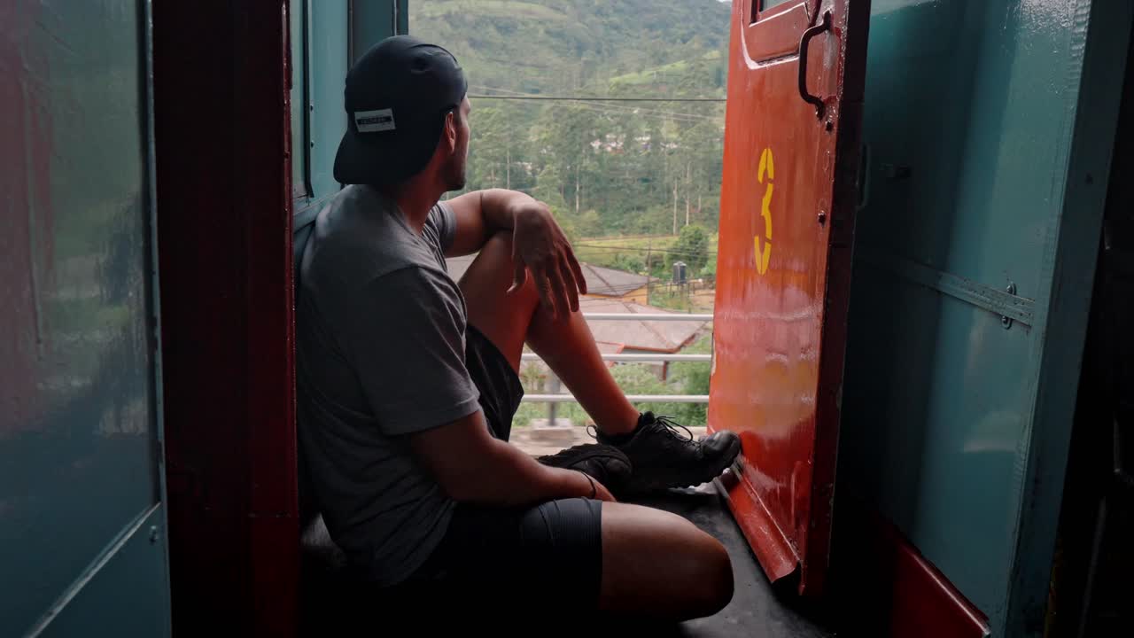 A man sits on the open door of a moving train along the scenic Ella to Kandy railway in Sri Lanka, gazing out at the lush green hills, tea plantations, and tropical jungle.