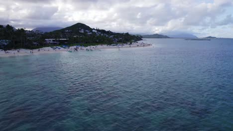 drone footage lanikai beach on the island of oahu showing reefs on the ocean floor with blue green water and a small beach town lining the coast and white sand beach, island mountains on the horizon