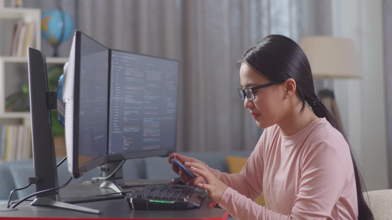 Asian Teen Girl Programmer Using Smartphone While Creating Software Engineer Developing App, Program, Video Game On Destop Computer At Home