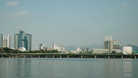 Hangang-waterfront-with-North-Seoul-cityscape-of-Mapo-gu,-Cars-Traffic-on-Gangbyeonbuk-ro-Expressway,-SEAH-Tower,-Mecenatpolis-Shopping-Mall,-Bukhansan-Mountains-on-background-on-sunny-day,-static