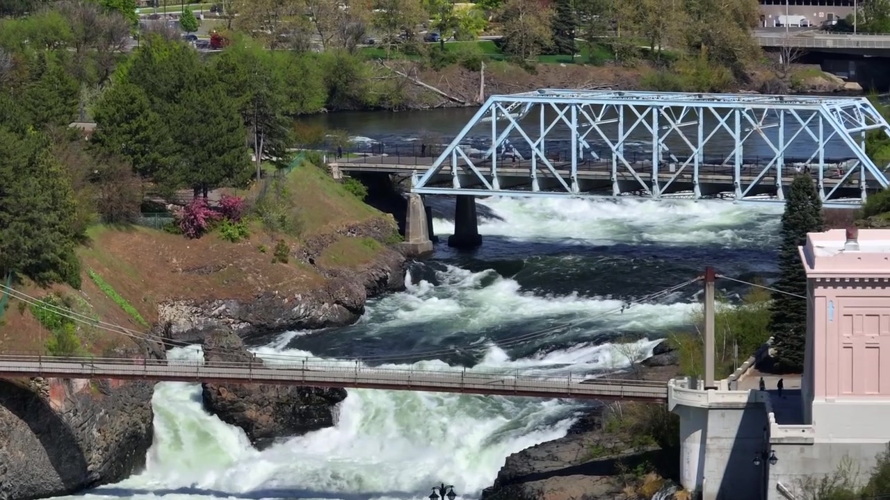 Spokane River Falls in Washington State with powerful rushing water and white splashes flowing under historic city bridges and pedestrian walkways during sunny day in USA. Aerial wide shot.