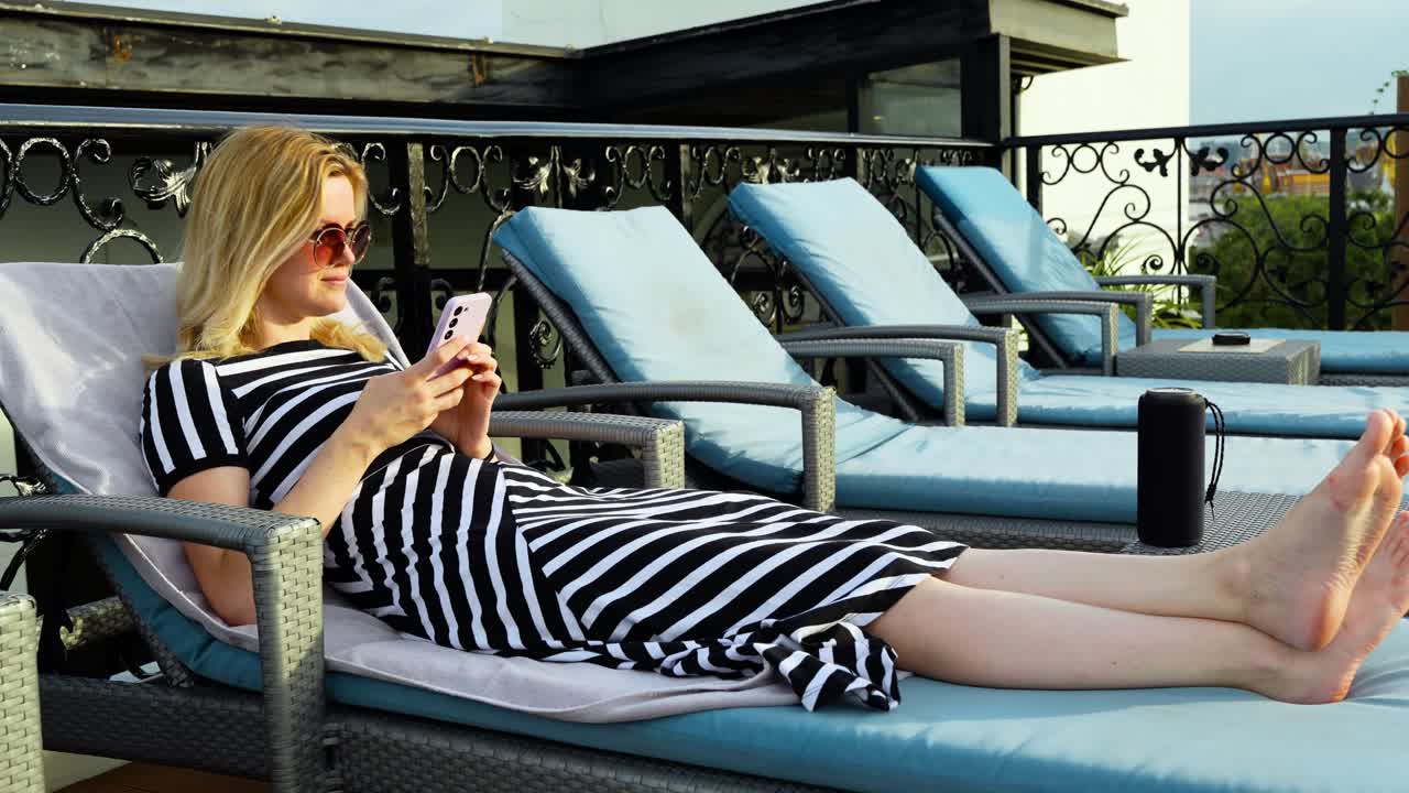 A woman relaxes on a rooftop lounger while using her phone and listening to music from a portable speaker, captured at a leisure resort type setting