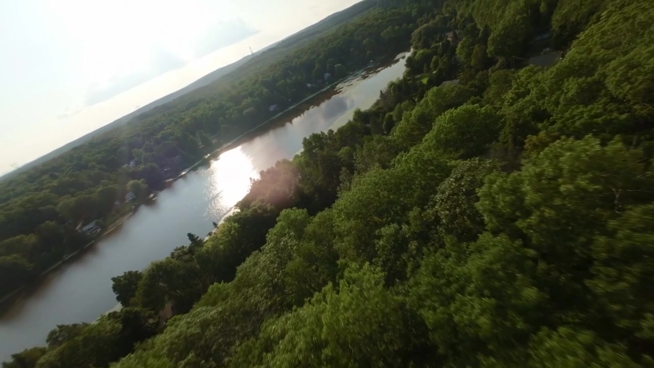 FPV drone flies over lush green trees and cabins near Siesta lake, Canada, with sunlit reflections on water.