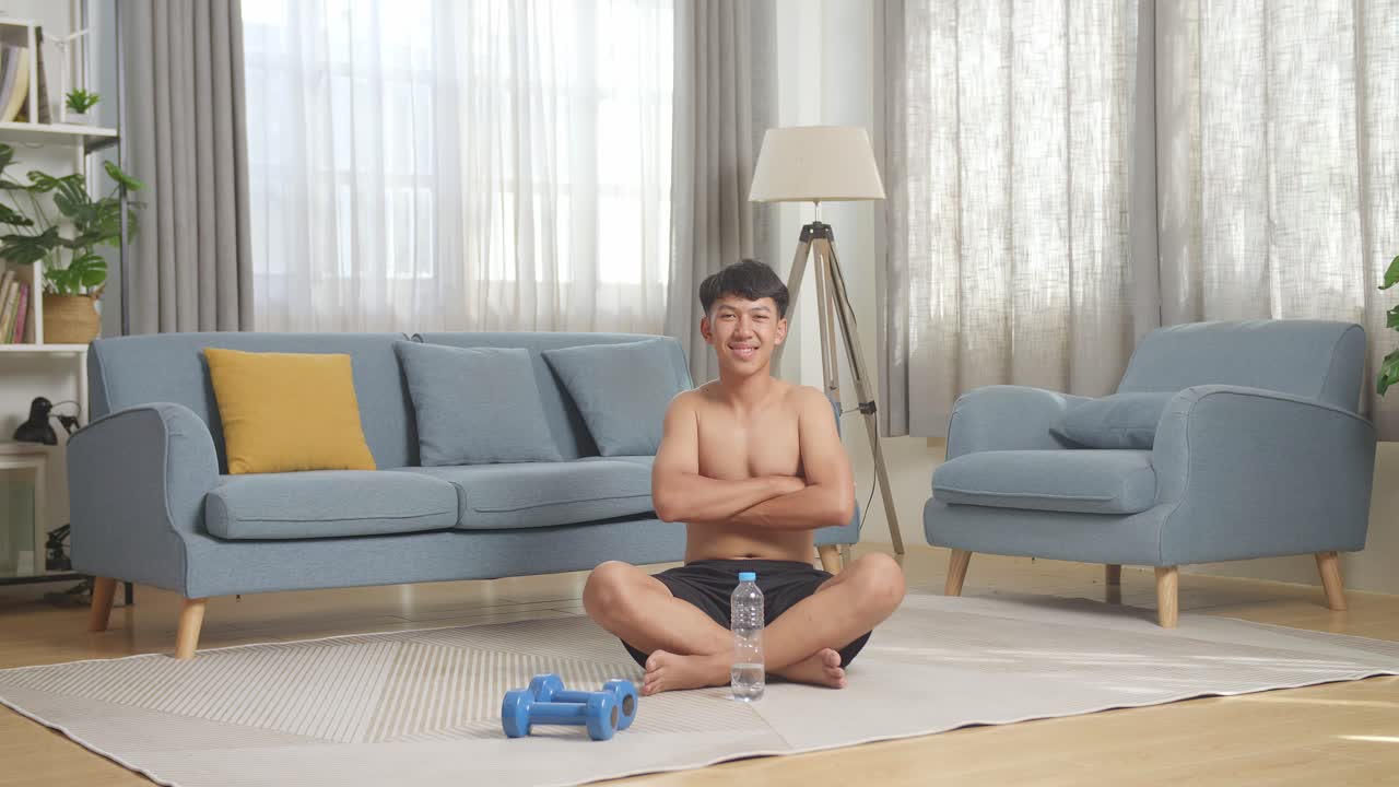 아시안 십대 소년의 몸이 가득 차 있으며, 집에서 셔츠를 벗고 운동한 후 덤벨과 함께 매트에 앉아 카메라를 향해 미소 짓고 있습니다.