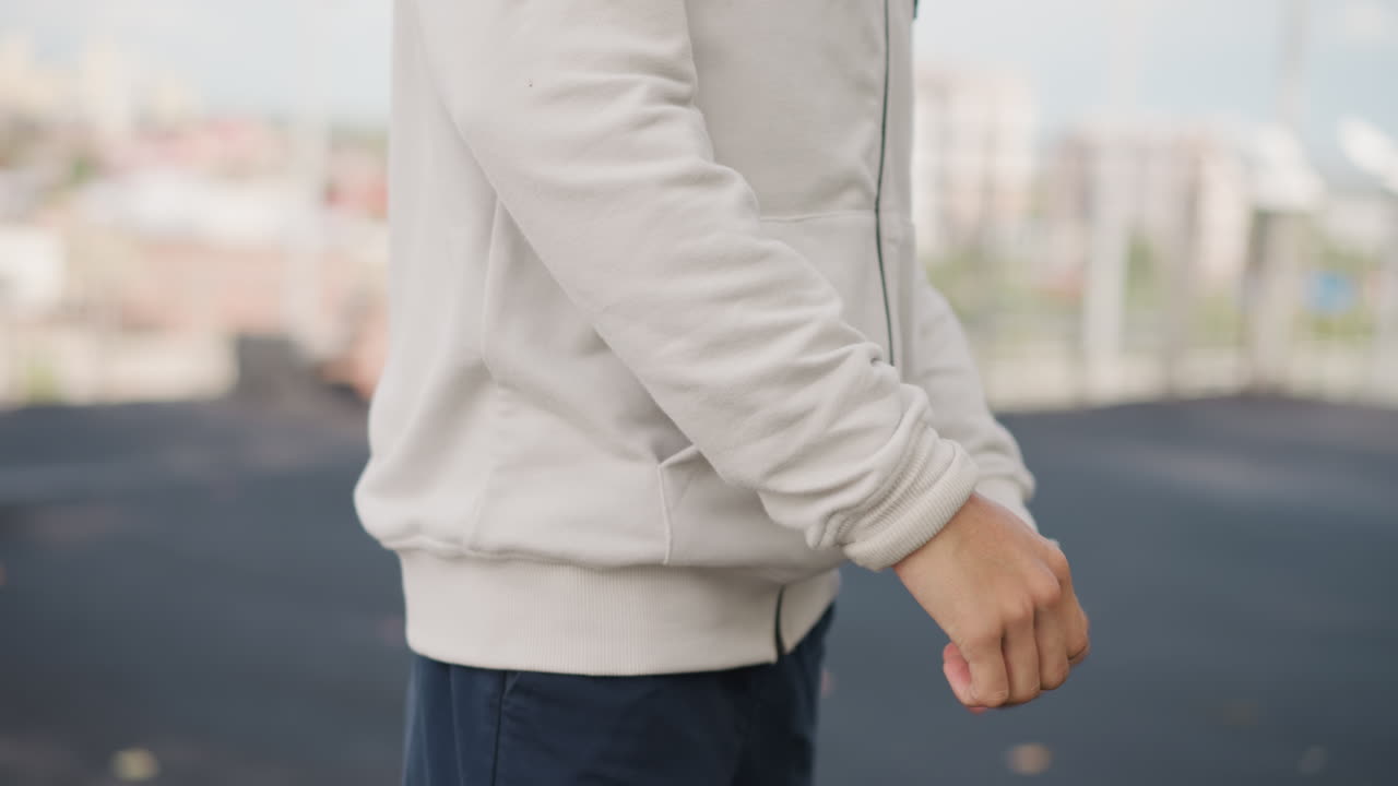 백인 남자가 옥상에서 소매를 걷고, 조깅 전 차분하게 후드티와 손목을 정리하며 준비 운동을 하고 있어요. 흐린 도시를 배경으로 조용하고 사색적인 분위기 속에서 집중하며 준비하는 모습이에요.