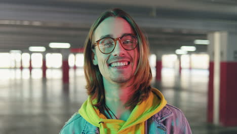 vista de cerca de un joven feliz y elegante con cabello largo y anteojos mirando a un lado y luego sonriendo a la cámara en un estacionamiento vacío