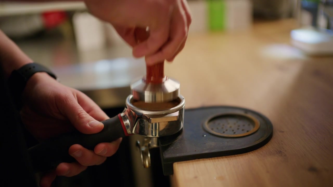 Close-up de um barista compactando pó de café com um tamper de metal dentro de um porta-filtro em um balcão de café de madeira