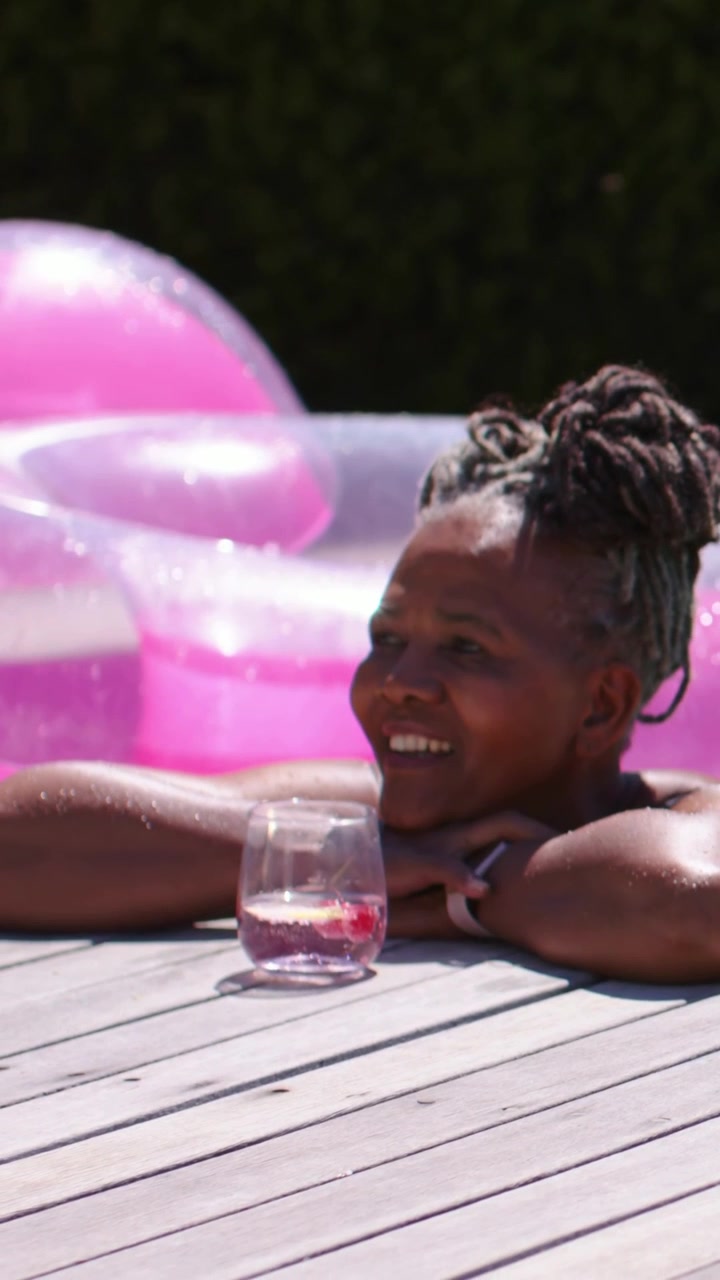 Vertical video: Leaning woman at pool edge, spotting person right, smiling with glass, straps