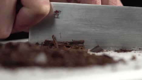 professional chef's hands chopping a dark chocolate bar using a kitchen knife on board, slow motion