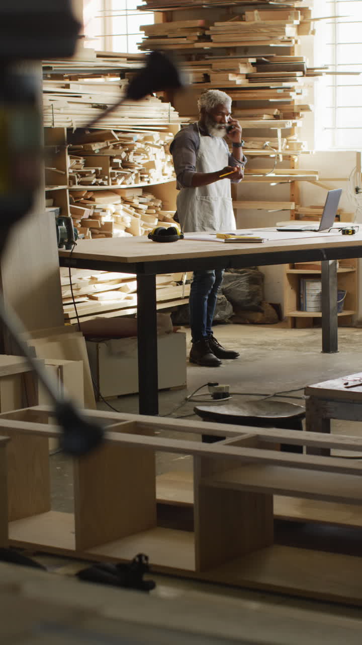 Vertical video of senior african american male carpenter using smartphone in workshop