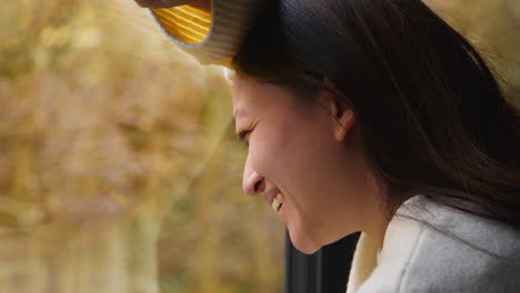 Close-Up-Of-Smiling-Woman-Leaning-Against-Window-At-Home-Looking-Out